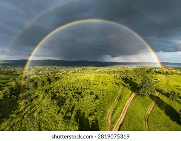 Dramatic storm sky with a vibrant full rainbow arch over green fields and rural landscape. Aerial drone photography.
