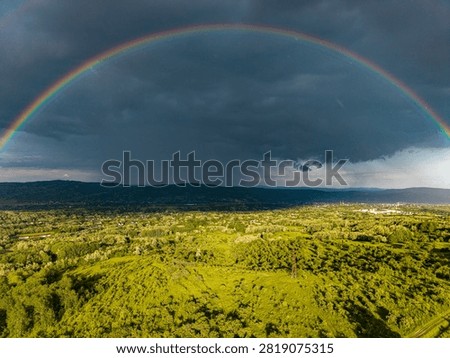 Dramatic storm sky with a vibrant full rainbow arch over green fields and rural landscape. Aerial drone photography.