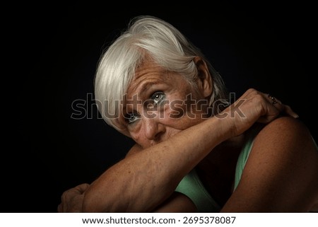 A dramatic studio portrait of an elderly woman resting her head on her crossed arms, gazing upward  The dark background and lighting, suggesting a mood of introspection or sadness.