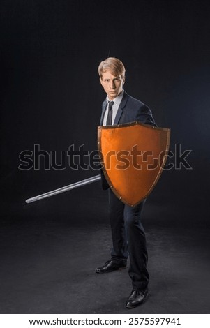In a dramatic studio pose, a man in a navy suit stands poised with a sword and shield. The dark background emphasizes the contrast between corporate and medieval imagery.
