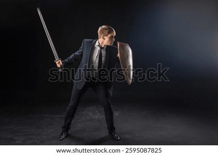 In a dramatic studio pose, a man in a navy suit stands poised with a sword and shield. The dark background emphasizes the contrast between corporate and medieval imagery.