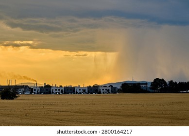 Dramatic summer thunderstorm with a heavy rain shaft pouring over a city skyline and wheat field at sunset.