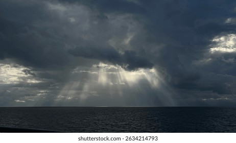 Dramatic sun rays pierce through dark storm clouds over the open ocean, creating a powerful and serene seascape