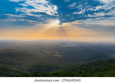 Dramatic sun rays pierce through clouds over a vast green forest and distant city at sunset in Rajasthan.