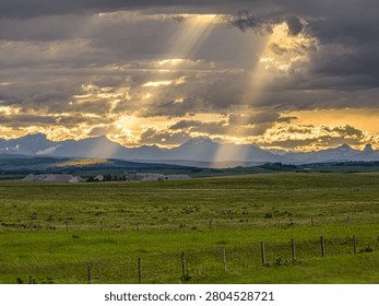 Dramatic sunbeams breaking through storm clouds over rolling green foothills and a distant mountain range during a golden hour sunset.