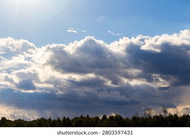 Dramatic sunbeams filtering through fluffy white cumulus clouds across a bright blue sky above a dark forest canopy during a scenic summer day with soft natural ambient lighting