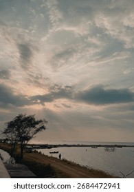 Dramatic sunbeams pierce through clouds over a calm sea during sunset. Silhouettes of a tree, boats, and people on the shore create a serene and hopeful coastal scene with a moody, natural ambiance.
