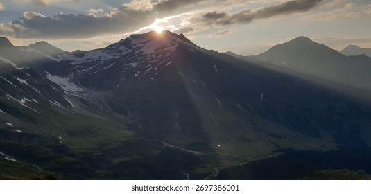 Dramatic sunburst as the setting sun dips behind a rugged mountain peak covered in snow. Intense light rays pierce the clouds and illuminate the deep green valley below.