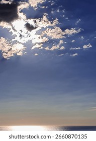 Dramatic sunlight breaking through clouds over a calm sea, creating a glowing reflection on the water below and a powerful contrast between light and sky.