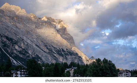 Dramatic sunlight breaking through rolling clouds over a rugged mountain peak above an alpine village in the Dolomites.