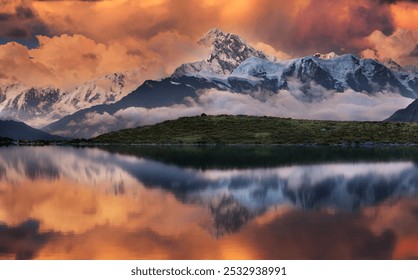 Dramatic sunlit clouds hover above snow-topped peaks, reflected in a serene lake with lush greenery between.