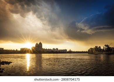 Dramatic sunset after a thunderstorm over Obolon Embankment in Kyiv, Ukraine. Golden sunbeams break through storm clouds, reflecting across the Dnipro River and city skyline.
