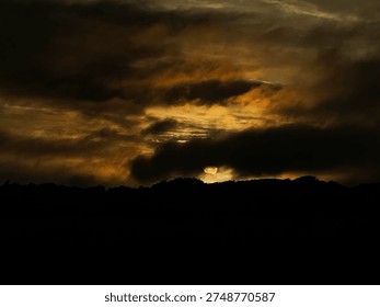 Dramatic Sunset Behind Dark Storm Clouds with Golden Amber Glow and Treeline Silhouette

A dramatic and atmospheric sunset with the sun disappearing behind a dark treeline silhouette, illuminating brooding storm clouds from below with rich golden and amber tones. Dark swirling storm clouds fill the frame creating a cinematic and mysterious evening sky scene.