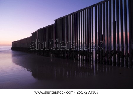 Dramatic sunset casting soft light over a towering border wall extending into the ocean, creating a stark contrast between natural beauty and man-made barriers.