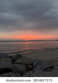 A dramatic sunset casts a brilliant red and orange glow over a calm sea, viewed from a sandy beach framed by large dark rocks in the foreground under heavy, dark storm clouds.
