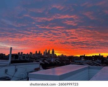 A dramatic sunset casts vibrant orange, red, and purple hues over the Philadelphia skyline, viewed from a residential rooftop in the Northern Liberties neighborhood. The city’s iconic buildings are si