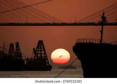A dramatic sunset casts a warm, fiery glow over an industrial harbor, silhouetting various vessels and structures against the vibrant sky. The large, round sun, a striking sphere of orange and red, dips towards the horizon, framed by the dark outlines of heavy-duty dredging ships or cranes in the midground. To the right, the majestic bow of a large ship with its railing and anchor chain creates a strong foreground element, hinting at the scale of maritime activity. In the background, the elegant cables of a suspension bridge stretch across the frame, adding architectural interest and depth to the urban coastal landscape. The tranquil water reflects the subdued light, while the hazy atmosphere adds a sense of timelessness and mystique to this bustling port scene. This powerful image captures the beauty of nature meeting human industry, perfect for themes of logistics, global trade, travel, infrastructure, and environmental subjects.