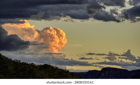 Dramatic sunset clouds glow above a rugged mountain landscape as dark storm clouds gather, contrasting warm golden light with brooding skies and conveying mood, power, and natural atmosphere.