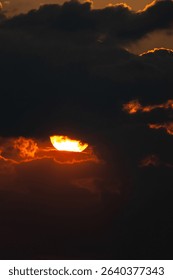 Dramatic sunset with glowing sun half-hidden behind thick dark clouds, casting a fiery glow on cloud edges and creating an intense, moody sky scene