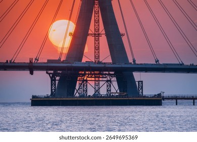 Dramatic sunset illuminating the under-construction Danshui cable bridge in New Taipei Taiwan, with the sun setting behind the towering structure over the water.