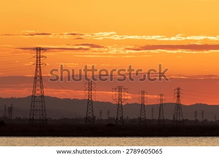 A dramatic sunset landscape featuring a line of high-voltage electrical transmission towers silhouetted against a brilliant orange and gold sky. Sunnyvale Bay Trail, Santa Clara County, California.