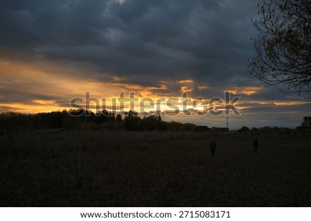 Dramatic sunset landscape with glowing clouds over a quiet path, dark silhouettes of people walking and trees in the distance, creating a moody and atmospheric evening scene.
