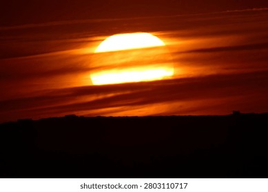 Dramatic sunset with the large sun partially obscured by thin horizontal clouds, casting intense orange and red hues across the sky.