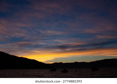 Dramatic sunset with orange and purple clouds over a dark mountain bay.
​High contrast twilight landscape showing the silhouette of mountains and small boats in a calm harbor. Vibrant sky with dramatic cloud formations reflected in the water. Perfect for travel, meditation, and inspirational content.