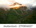 Dramatic sunset over the abandoned mountain settlement of Invernales del Texu in the Picos de Europa, Asturias, Spain. Low clouds and mist wrap around rugged limestone peaks while golden morning light illuminates the lush green valley, traditional shepherd huts, and winding mountain road, creating a breathtaking landscape scene in northern Spain.