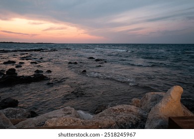 Dramatic sunset over the Adriatic Sea in Puglia. Golden-orange sky contrasts with dark clouds and deep blue waters. Rocky shoreline and silhouetted figure create a moody, contemplative coastal scene. - Powered by Shutterstock