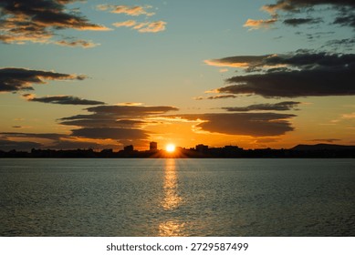 a dramatic sunset over a calm body of water, with the sun bursting through clouds above a silhouetted city skyline and casting a golden reflection on the ripples below.
