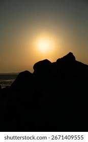 A dramatic sunset over a calm ocean, with the sun's golden light illuminating the hazy sky above the dark silhouettes of rocky formations in the foreground.