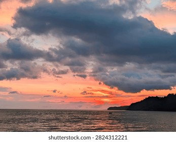 ​Stunning dramatic sunset over the calm ocean with vibrant orange and dark blue clouds filling the sky, featuring a silhouette of a coastal hill and gentle sea waves.