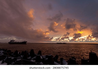 Dramatic sunset over calm sea with dark clouds, glowing orange sky, ship silhouette and harbor foreground, creating a moody coastal evening scene