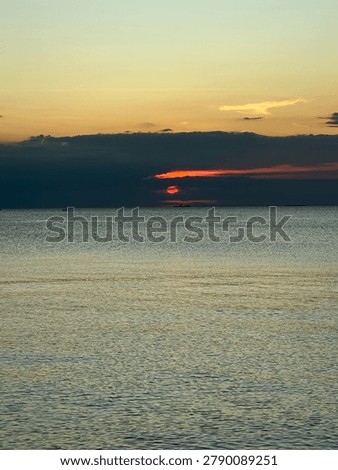 Dramatic sunset over the calm sea with a bright red sun hiding behind dark storm clouds.