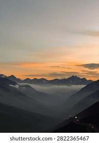 Dramatic sunset over a deep mountain valley filled with thick fog, featuring silhouetted mountain peaks and a colorful orange sky.