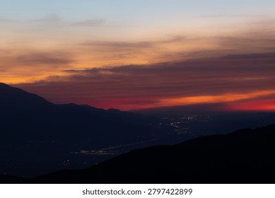 Dramatic sunset over a desert valley with silhouetted mountains in the foreground and scattered town lights glowing below as twilight approaches.