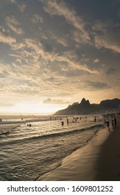 A Dramatic Sunset Over Ipanema Beach In Rio De Janeiro, Brazil,with Golden Light From The Low Angle Sun Shining Up Onto The Stripes Of Grey Clouds. There Are Silhouettes Of People Swimming In The Sea.