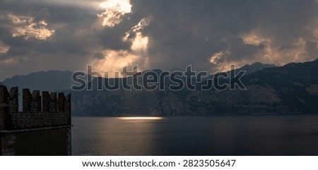 A dramatic sunset over lake, with mountains in the background and castle foreground. Sunbeams pierce through clouds, casting warm glow on water mountains.