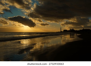 Dramatic sunset over Maspalomas beach in Gran Canaria with the iconic lighthouse silhouette. Golden sunlight reflects on the wet sand and Atlantic waves under a cloudy sky at twilight