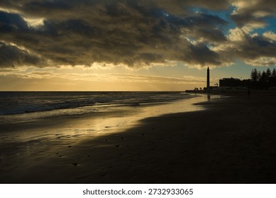 Dramatic sunset over Maspalomas beach in Gran Canaria with the iconic lighthouse silhouette. Golden sunlight reflects on the wet sand and Atlantic waves under a cloudy sky at twilight