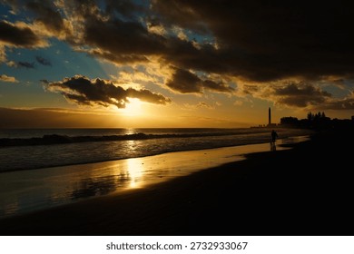 Dramatic sunset over Maspalomas beach in Gran Canaria with the iconic lighthouse silhouette. Golden sunlight reflects on the wet sand and Atlantic waves under a cloudy sky at twilight