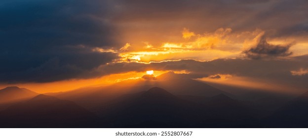 Dramatic sunset over misty mountain peaks, with golden light piercing through dark, ominous clouds. Awe-inspiring nature scene with a sense of power and wonder. Perfect for travel, nature, or spiritua