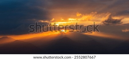 Dramatic sunset over misty mountain peaks, with golden light piercing through dark, ominous clouds. Awe-inspiring nature scene with a sense of power and wonder. Perfect for travel, nature, or spiritua