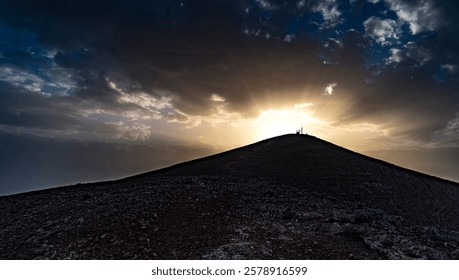 A dramatic sunset over a mountain peak, with rays of sunlight breaking through dark clouds. Silhouetted figures stand on the summit, creating a striking contrast against the vibrant sky.