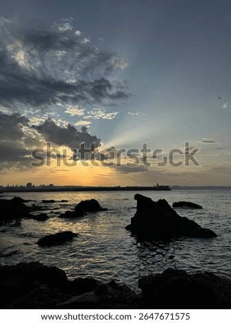 A dramatic sunset over the North Sea as seen from the Torry Battery in Aberdeen, Scotland. Jagged rocks silhouette against the golden water, while the city skyline and distant lighthouse are hazy. 