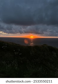 dramatic sunset over the ocean. Dark clouds create a sense of tension, while the fiery orange sun breaks through, casting a glow on the calm water. In the foreground, a grassy cliff with white flowers