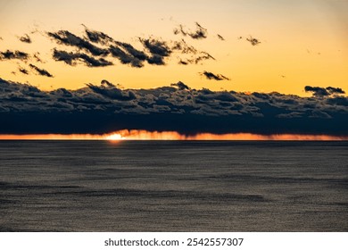 A dramatic sunset over the ocean, with dark clouds partially covering the horizon. Fiery orange and yellow hues from the setting sun illuminate the edges of the clouds, contrasting with the calm sea.