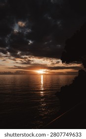 Dramatic sunset over the ocean with dark storm clouds, golden sunlight reflecting across calm water, and a peaceful coastal horizon at dusk.
