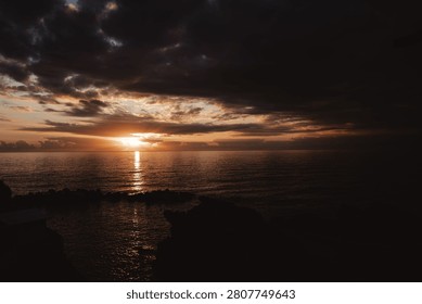 Dramatic sunset over the ocean with dark storm clouds, golden sunlight reflecting across calm water, and a peaceful coastal horizon at dusk.